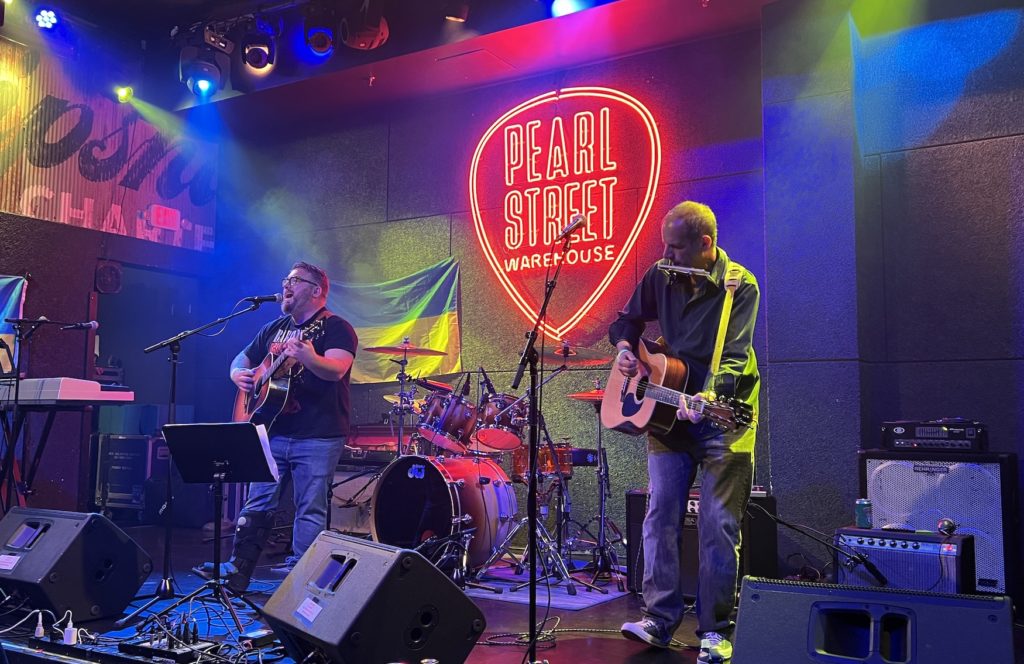 Seán O'Connor and Jon Knight rock out on the Pearl Street Warehouse stage under blue and yellow lights and in front the the Ukrainian flag